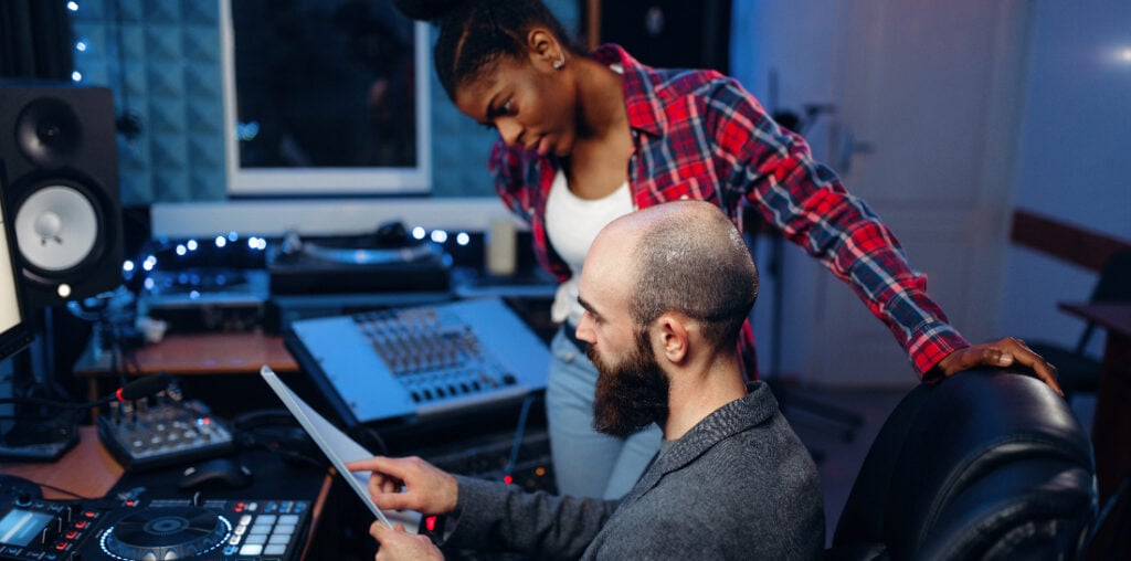 Bearded sound engineer at remote control panel in audio recording studio talking with a woman