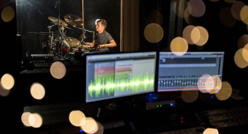 A drummer in a recording studio as seen through the control room glass with recording equipment to illustrate Why Do So Many Musicians Live in LA?